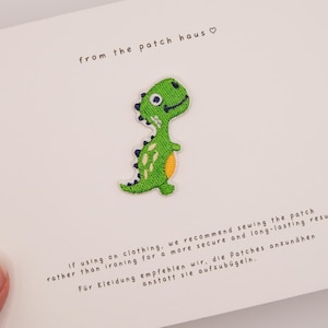 Op de afbeelding: Een geborduurde groene dinosauruspatch met een gele buik en donkerblauwe stekels, gecentreerd op een witte kaart. De kaart bevat de tekst from the patch haus en instructies om de patch op kleding te naaien voor een duurzaam resultaat.