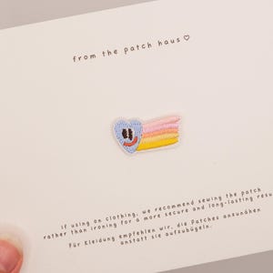 Op de afbeelding: Een geborduurde hartvormige patch met een lichtblauw gezicht, bruine ogen en een oranje glimlach, gevolgd door een roze, gele en oranje regenboog. De patch is bevestigd op een witte kaart bedrukt met de tekst from the patch haus en instructies.