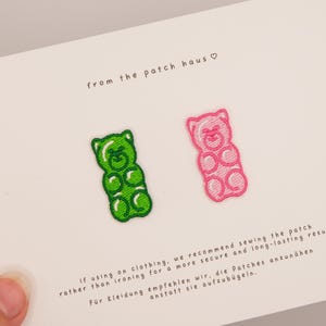Könnte beinhalten: Zwei gestickte Gummibärchen-Aufnäher, einer grün und einer pink, auf einer weißen Karte. Die Karte enthält den Text from the patch haus und Anweisungen, die empfehlen, die Aufnäher für ein sicheres und langlebiges Ergebnis auf Kleidung zu nähen.