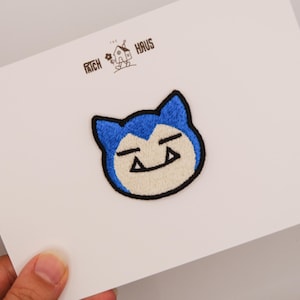Könnte beinhalten: Ein gestickter Aufnäher mit dem Gesicht einer blau-cremefarbenen Kreatur mit spitzen Ohren und schwarzer Umrandung. Der Aufnäher ist auf einer weißen Karte befestigt, auf der oben der Text THE PATCH HAUS und eine kleine Hausillustration gedruckt sind.