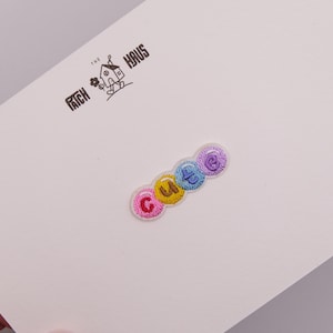 May include: An embroidered patch featuring the word CUTE in individual circular segments with pink, yellow, blue, and purple thread colors. The patch is attached to a white card printed with the black logo THE PATCH HAUS featuring a small house icon.