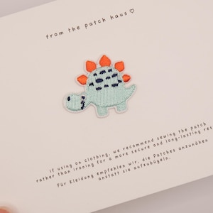 May include: An embroidered dinosaur patch featuring a light blue body with dark blue spots and orange dorsal plates. The patch is attached to a white card with the text from the patch haus and instructions for sewing the patch onto clothing.
