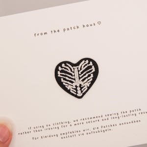 Könnte beinhalten: Ein schwarzer herzförmiger Aufnäher mit einem weißen gestickten Brustkorb-Motiv. Der Aufnäher ist auf einer weißen Karte befestigt, die den Text from the patch haus und Hinweise zum Annähen des Patches an Kleidung enthält.