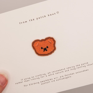 May include: An embroidered iron-on patch featuring a small, brown bear face with black stitched eyes and a nose. The patch is centered on a white display card with the text from the patch haus and instructions for sewing the patch onto clothing.