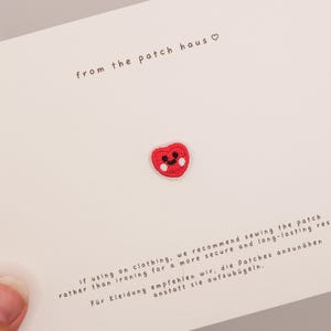Könnte beinhalten: Ein kleiner roter, herzförmiger Stickaufnäher mit einem lächelnden Gesicht, schwarzen Augen und weißen Wangen. Der Aufnäher ist auf einer weißen Karte befestigt, die den Text from the patch haus sowie Anweisungen zum Annähen enthält.