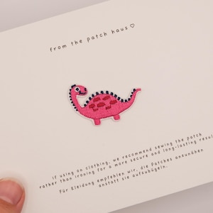 Könnte beinhalten: Ein gestickter Dinosaurier-Aufnäher in leuchtendem Pink mit dunkelblauen Akzenten auf einer weißen Karte. Die Karte enthält den Text from the patch haus sowie Hinweise, den Aufnäher für eine längere Haltbarkeit aufzunähen.