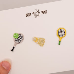 Può includere: Tre toppe ricamate su un cartoncino bianco con la scritta THE PATCH HAUS. Le toppe includono una racchetta da tennis con una pallina verde, un volano da badminton giallo e bianco e una racchetta da tennis gialla con impugnatura verde.