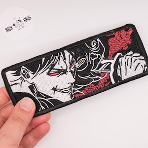 May include: A rectangular embroidered patch featuring a black and white anime-style character with red eyes and a sharp grin, holding a sword. The design includes red accents and a black border. The patch is held against a plain white background.