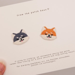 Puede incluir: Dos parches bordados termoadhesivos con forma de caras de perro Shiba Inu, uno en blanco y negro y otro en naranja y blanco, presentados sobre una tarjeta blanca con el texto from the patch haus e instrucciones para coser los parches a la ropa.