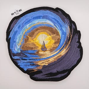 Könnte beinhalten: Ein kreisförmiger bestickter Aufnäher mit einem Segelboot auf dem Wasser bei Sonnenuntergang. Das Design zeigt eine leuchtend gelbe Sonne, wirbelnde blaue und violette Wolken sowie dunkle Umrisse. Der Text PATCH HAUS ist oben links zu sehen.