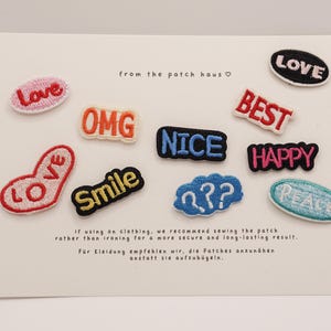 Könnte beinhalten: Eine Sammlung von neun gestickten Stoffaufnähern auf einer weißen Präsentationskarte. Die Aufnäher zeigen verschiedene Wörter und Symbole wie Love, OMG, Best, Nice, Happy, Smile, Peace und eine blaue Wolke mit weißen Fragezeichen.