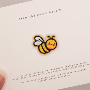 Puede incluir: Un parche bordado que muestra una abeja amarilla y negra con alas blancas y una cara sonriente. El parche está sujeto a una tarjeta blanca con el texto from the patch haus e instrucciones para coser el parche en prendas de vestir.