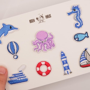 Könnte beinhalten: Eine Sammlung von neun nautischen Stickaufnähern auf einer weißen Karte, darunter ein Delfin, Seepferdchen, Oktopus, Fisch, Heißluftballon, Kreuzfahrtschiff, Rettungsring, Leuchtturm und Segelboot. Der Text THE PATCH HAUS ist in Schwarz aufgedruckt.
