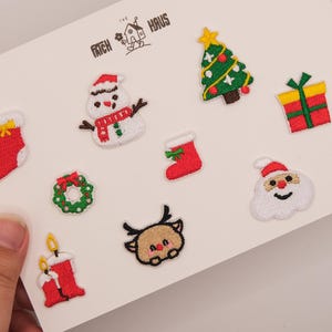 Puede incluir: Un conjunto de diez parches bordados con motivos navideños sobre una tarjeta blanca con la etiqueta The Patch Haus. Los diseños incluyen un calcetín rojo, un muñeco de nieve, un árbol verde, un regalo, una corona y a Santa Claus.