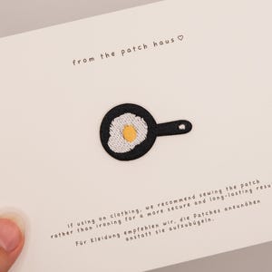 May include: An embroidered iron-on patch shaped like a black frying pan with a white egg and yellow yolk, centered on a white card. The card features the text 'from the patch haus' and instructions in English and German regarding application.