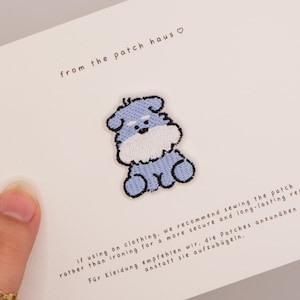 Puede incluir: Un parche bordado de un perro azul claro y blanco con un contorno oscuro, centrado en una tarjeta blanca. La tarjeta presenta el texto from the patch haus, junto con instrucciones en inglés y alemán sobre cómo coser el parche en la ropa.
