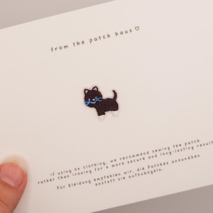 Può includere: Una piccola toppa ricamata raffigurante un gatto nero con zampe bianche e un collare blu, centrata su un cartoncino bianco. Il cartoncino riporta la scritta 'from the patch haus' e istruzioni in inglese e tedesco su come applicare la toppa.