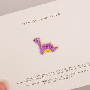 Puede incluir: Un pequeño parche bordado de un dinosaurio morado con púas naranjas y vientre amarillo, presentado sobre una tarjeta blanca con el texto from the patch haus e instrucciones para coser el parche en prendas de vestir.