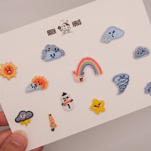 Può includere: Una collezione di piccole toppe ricamate su un cartoncino bianco con il logo "THE PATCH HAUS". I disegni includono un sole sorridente, un arcobaleno, un pupazzo di neve, una stella gialla, varie nuvole blu con faccine, un tornado e un piccolo razzo arancione.