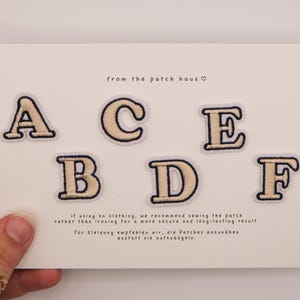 Op de afbeelding: Zes geborduurde alfabet-patches met de letters A, B, C, D, E en F in een crèmekleur met donkerblauwe randen. De patches worden getoond op een witte kaart met de tekst from the patch haus en instructies voor het naaien.