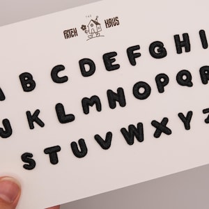Puede incluir: Un conjunto de parches bordados con las letras del alfabeto en color negro sobre una tarjeta blanca. Las letras de la A a la Z se muestran en una fuente negrita y redondeada. Arriba, en el centro, un logo muestra una casa y el texto THE PATCH HAUS.