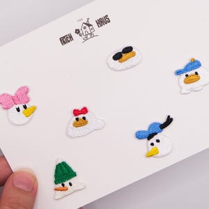 Op de afbeelding: Een set van zes geborduurde strijkpatches met cartoon-eendengezichten en diverse accessoires, waaronder een roze strik, een groene muts, een zonnebril en blauwe petten, gepresenteerd op een witte kaart met het logo van The Patch Haus.