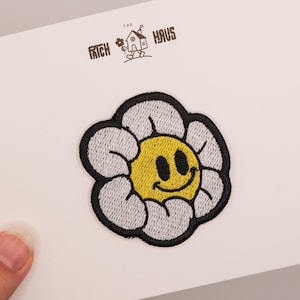 Könnte beinhalten: Ein gestickter Aufnäher in Form einer weißen Blume mit gelber Mitte und einem schwarzen Smiley-Gesicht. Der Aufnäher hat einen dicken schwarzen Rand. Darüber steht der Text THE PATCH HAUS mit einer kleinen Abbildung eines Hauses mit Blume.