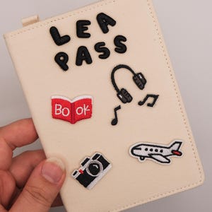 May include: A cream-colored passport holder featuring black embroidered text that reads LEA PASS, along with several colorful patches including a red book, black headphones with musical notes, a black and white camera, and a white airplane.