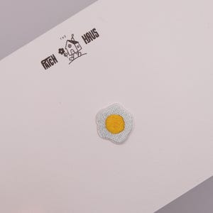 Puede incluir: Un parche bordado con forma de huevo frito con una yema amarilla brillante y bordes blancos, centrado en una tarjeta blanca. La tarjeta presenta el texto impreso en negro THE PATCH HAUS con un pequeño gráfico de una casa con una flor.