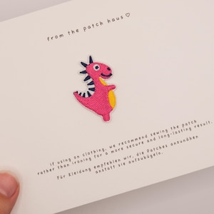 May include: An embroidered pink dinosaur patch with a yellow belly and black and white striped back spikes. The patch is attached to a white card featuring the text from the patch haus and instructions for sewing the patch onto clothing.