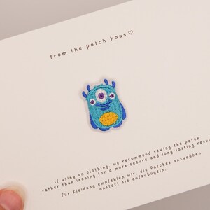 May include: An embroidered patch featuring a blue monster with three eyes, a wide smile, and a yellow oval belly. The patch is centred on a white card with the text from the patch haus and instructions for sewing the patch onto clothing.