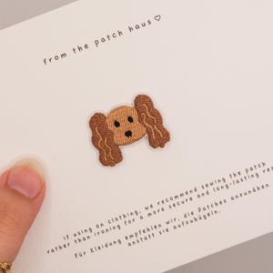 May include: An embroidered iron-on patch featuring a tan dog face with long, wavy brown ears and black stitched eyes and nose. The patch is centered on a white card with the text from the patch haus and sewing instructions in English and German.