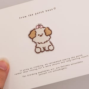 Puede incluir: Un parche bordado de un pequeño cachorro blanco con orejas marrones y un lazo rosa en la cabeza, centrado en una tarjeta blanca. La tarjeta presenta el texto from the patch haus e instrucciones para coser el parche en la ropa.