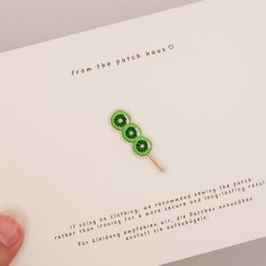 Könnte beinhalten: Ein gestickter Aufnäher in Form eines Spießes mit drei grünen Kiwischeiben an einem hellen Holzstäbchen, zentriert auf einer weißen Karte mit dem Text from the patch haus und Anweisungen zum Annähen des Aufnähers an Kleidung.