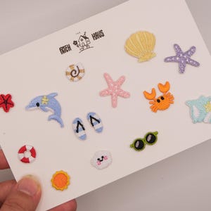 Peut inclure: Un ensemble de petits écussons brodés sur une carte blanche portant le texte THE PATCH HAUS. Les motifs incluent un coquillage jaune, des étoiles de mer violettes et roses, un crabe orange, un dauphin bleu, une bouée rouge et blanche, des tongs, des lunettes de soleil et un soleil.