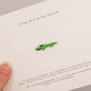 May include: A small green embroidered crocodile patch centred on a white display card. The card features the text from the patch haus with a heart symbol, along with instructions in English and German regarding sewing the patch onto clothing.