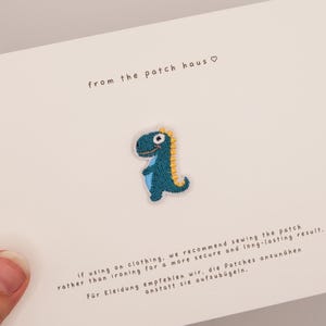 May include: A small embroidered dinosaur patch featuring a teal body, light blue belly, and yellow spikes. The patch is centered on a white card with the text from the patch haus and instructions for sewing the patch onto clothing items.