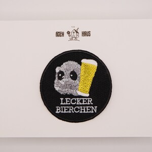 May include: A circular embroidered patch featuring a grey furry creature holding a yellow beer glass, with the text LECKER BIERCHEN in white below. Above the patch, the text THE PATCH HAUS is printed with a small house icon in the centre.