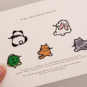 Pode incluir: Um conjunto de cinco emblemas bordados de animais num cartão branco, incluindo um panda, um coelho, uma tartaruga, um gato malhado laranja e um gato cinzento. O texto from the patch haus está impresso no topo com instruções de aplicação.