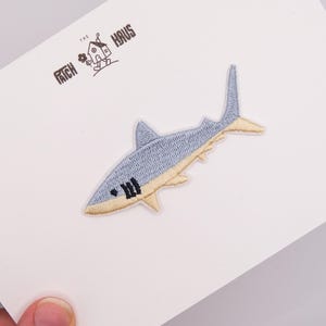 May include: An embroidered shark patch featuring a light blue top and a cream-colored underside, displayed on a white card with the text THE PATCH HAUS and a small house illustration printed in black ink at the top left corner of the card.