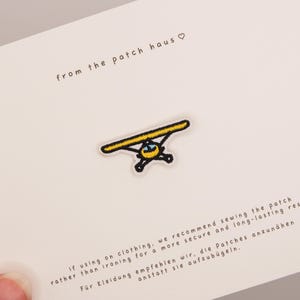 May include: An embroidered patch featuring a small yellow airplane with black outlines and a blue cockpit window. The patch is centered on a white card with the text from the patch haus and instructions for sewing the patch onto clothing.