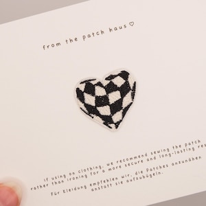 Könnte beinhalten: Ein herzförmiger bestickter Aufnäher mit schwarz-weißem Karomuster, zentriert auf einer weißen Karte. Die Karte enthält den Text from the patch haus sowie Anweisungen auf Englisch und Deutsch zum Annähen des Aufnähers auf Kleidung.