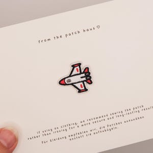 May include: An embroidered iron-on patch featuring a white, red, and black airplane design. The patch is centered on a white card with the text from the patch haus and instructions for sewing the patch onto clothing for a secure result.