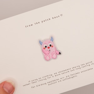 Op de afbeelding: Een geborduurde patch met een klein roze monster met blauwe hoorns, rode wangen en een zwarte neus, gecentreerd op een witte kaart. De kaart bevat de tekst "from the patch haus" en instructies voor het bevestigen van de patch op kleding.
