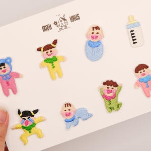 May include: A set of eight embroidered iron-on patches on a white backing card labeled The Patch Haus. The designs include various babies in colorful outfits, a swaddled infant, and a baby bottle with a yellow cap and blue base.