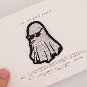 Può includere: Una toppa ricamata raffigurante un fantasma bianco che indossa occhiali da sole neri e tiene una sigaretta in bocca. La toppa è fissata su un cartoncino bianco con il testo from the patch haus e le istruzioni per il cucito stampate.