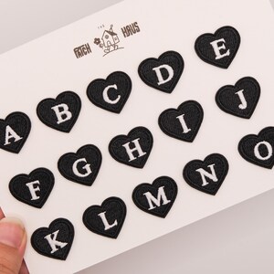 Puede incluir: Una colección de quince parches bordados en forma de corazón negro, cada uno con una letra mayúscula blanca de la A a la O. Los parches están dispuestos sobre una tarjeta blanca que muestra el texto THE PATCH HAUS con un dibujo de una casa.