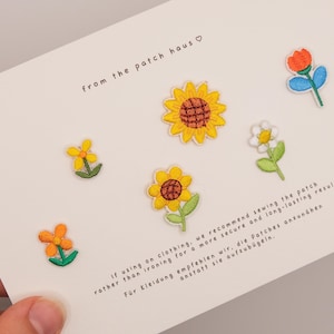 Könnte beinhalten: Ein Set aus sechs gestickten Blumenaufnähern auf einer weißen Karte. Die Aufnäher zeigen eine große gelbe Sonnenblume, eine kleinere gelbe Blume, ein weißes Gänseblümchen, eine orangefarbene Blume und eine rote Tulpe. Text from the patch haus.