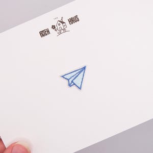 Könnte beinhalten: Ein kleiner gestickter Aufnäher in Form eines Papierfliegers mit hellblauen und dunkelblauen Faden-Details. Der Aufnäher ist auf einer weißen Karte zentriert, die den schwarzen Aufdruck THE PATCH HAUS und eine kleine Haus-Illustration zeigt.