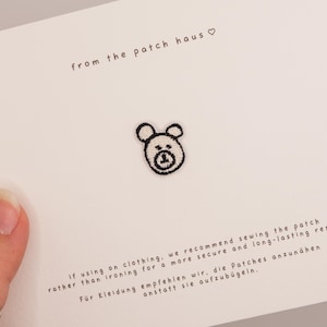 May include: A small black embroidered bear face patch centered on a white card. The card features the text from the patch haus with a heart symbol, and instructions recommending sewing the patch onto clothing for a secure and long-lasting result.